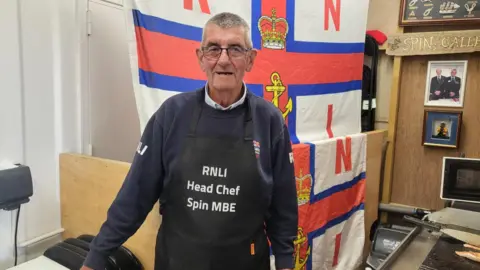 Richard Spindler Richard standing in an RNLI office wearing an apron that says "RNLI head chef Spin MBE". He is wearing glasses and has short grey hair.