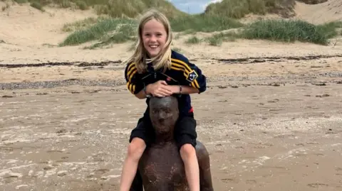 A boy with long blond hair sits on the shoulders of a statue on a beach and smiles at the camera. 