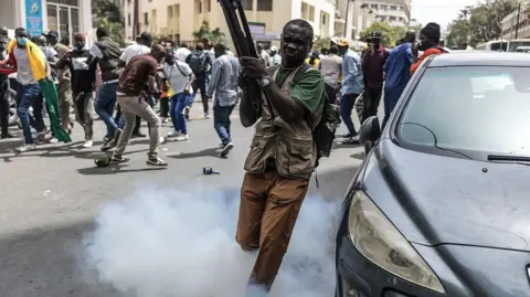 A man carrying a tripod escapes from a tear gas as smoke fills a street, while protesters scatter and gather in the background during clashes 
