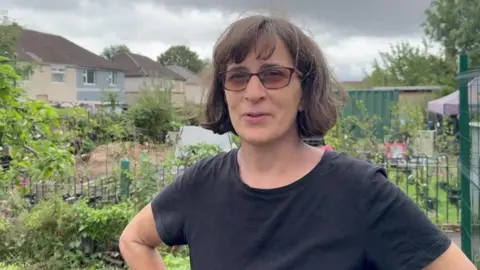 Samantha Turner has mid length brown hair and is wearing brown framed sunglasses and a black t-shirt. She is stood in a garden with green plant sin the bacvkground.