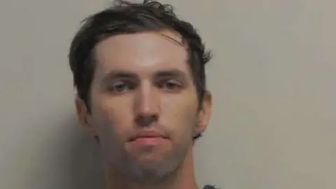 Utah Governor's office A young white man with dark short hair swept to the side, light eyes, and noticeable stubble on his chin and above his lip stares unsmiling into the camera