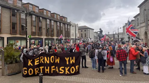 Crowds of people in Truro city centre with Union Jack flags and signs. There are buildings either side, and a statue towards the back. On the left, there are people holding a large black sign which has the words 'KERNOW YW GORTHFASKOR' in yellow writing.