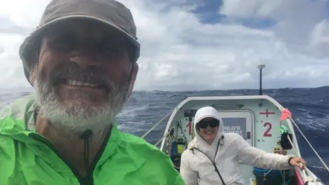 Nicky and Bob Parr are in their boat on the ocean. Mr Parr has a beard and is wearing a green jacket and hat. His wife is wearing a white, hooded jacket and sunglasses.