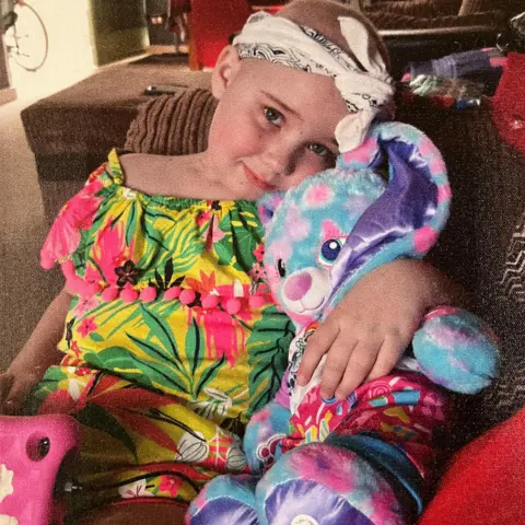 Mark Bissett Charly in hospital aged seven as a cancer patient - A child wearing a brightly coloured floral outfit sits on a couch while holding a plush toy rabbit with multicoloured fur and long ears.