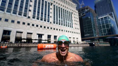 A swimmer wearing goggles and a green hat with his body up to his shoulders is in the water of the swimming venue in Canary Wharf. Tall glass-fronted buildings are in the background. 