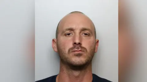 A police mugshot of Sean Deering - a man in his 30s with his shaved hair and stubble. He has brown eyes and is looking into the camera.