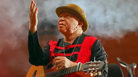 Salif Keita wearing a black and re shirt and ochre bowler hat holds his right hand in the air as he looks upwards. A guitar is resting on his knee and smoke can be seen behind him.