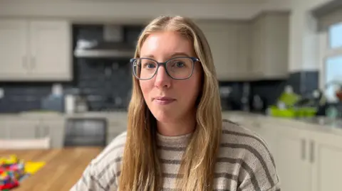 Laura Magee has long dark blonde hair and is wearing square glasses. She is looking down the camera lens with her lips pursed. She is sitting at a wooden table in a white and black kitchen.