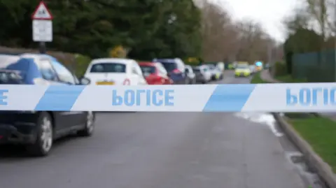 Blue and white police tape indicates a cordon on the road, which is lined by parked cars. A police car and an ambulance are in the background. The road is lined by trees and bushes.