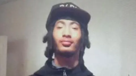 Kalvin Taylor looks directly at the camera with a serious expression. He is wearing a black zip-up hoodie and a black cap.