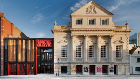 Philip Vile The frontage of the Bristol Old Vic theatre. It is a large historic sand coloured building with tall sash windows and large pillars. On the left there is a more modern addition to the building, with black and red metal beams and a large sign saying 'Bristol Old Vic'. 