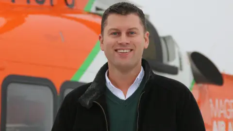 Magpas Daryl Brown, smiling and looking at the camera, by a helicopter, wearing a black jacket, green jumper and white shirt, he has short dark hair.