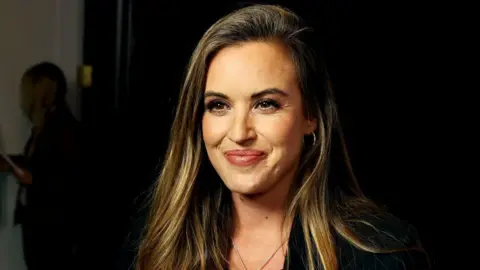 Charlie Webster smiles at the camera. She has long brow hair and is wearing a black jacket and top.