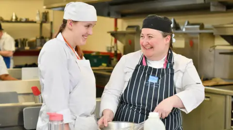 A picture of Abigail and her lecturer in a college kitchen. They are both smiling at each other. They are both wearing a white outfit with an apron on top. They are both wearing a hat