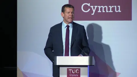 BBC Rhun ap Iorwerth speaking at a lectern, wearing a dark suit, blue shirt and a dark purple tie