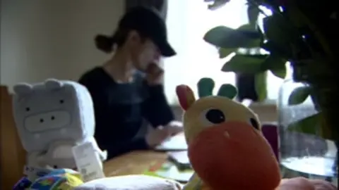 BBC A woman wearing a dark baseball cap and T-shirt sits in front of a window with children's toys in the foreground