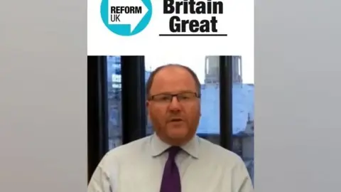 Facebook This is a snapshot taken from the deepfake video. A man who appears to be George Freeman is sitting in front of a window, talking directly to the camera. He is wearing a shirt and tie, and glasses. Above him is the Reform Party logo and the words "Reform UK - Make Britain Great"