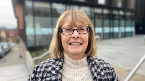 Laura Coffey/ BBC Wendy Randall, who has dark blonde hair and glasses, smiles as she looks at the camera. She is wearing a cream cable jumper and a black-and-white knitted cardigan. Behind her, and out of focus, is what looks like an office building.