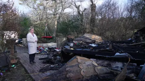 Woman with her hair tied up in a pony-tail on top of head and wearing a cream pullover ... looking at her burnt out caravan 