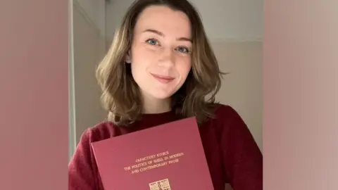 Supplied Dr Ally Louks is holding her thesis - a red A4 sized hardback book. Its title is written in gold lettering on the front. Dr Louks is smiling at the camera. She has shoulder length light brown hair, blue eyes and is wearing a burgundy jumper. 