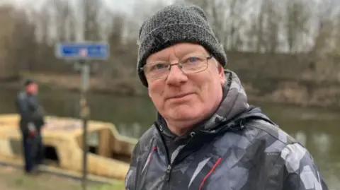 BBC/Nicola Rees Ian Platt stands on the banks of the River Aire with a small boat in the background. He is wearing a woolen cap, waterproof jacket and glasses.