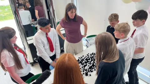 Seven young secondary school pupils and a teacher standing in front of desk which has some black and white balls on it which represent the HPV virus