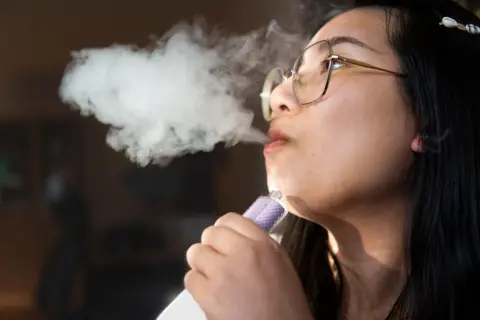 A young person exhaling smoke from a vape, which she is holding in her hand. 