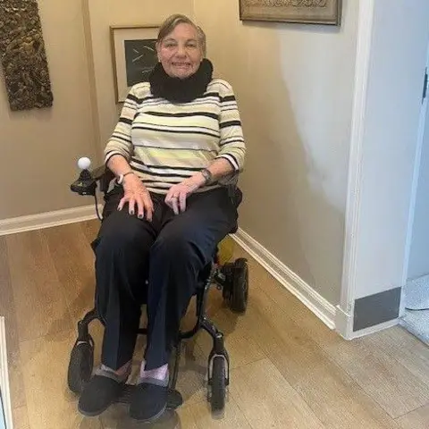 Joyce Williams A woman sitting in a wheelchair in a hallway. She is wearing a a striped jumper with a black scarf around her neck.