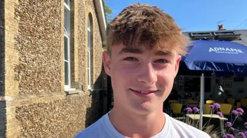 Vikki Irwin/BBC Archie smiling at the camera, wearing a white T-shirt stood in front of a stone building with a blue umbrella in the background.