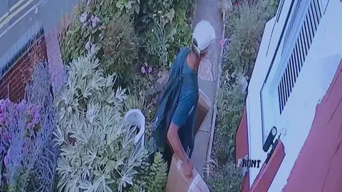 A man walks away from the front door of a house with two brown parcels in his hands. He has a cap on and bad on his back as he walks on a short garden path through a front garden away form a house.