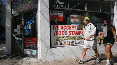 Getty Images Two people walking past a small corner store with a sign that says it accepts food stamps