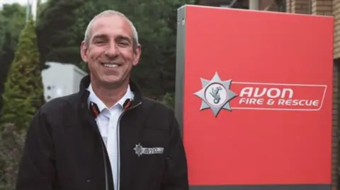 Avon Fire & Rescue Service Simon Shilton is pictured smiling at the camera, wearing an Avon Fire and Rescue fleece, next to a large sign for the organisation. He has short dark hair, and is standing outside what appears to be the service's headquarters. 