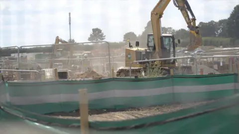 The image shows a construction site where two excavators can be seen, as well as a lot of dust in the air.