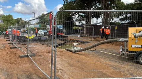 An area of road is cordoned off by construction fencing as heavy machinery gets to work and construction workers work around it. A pipe is extended from a tanker into a large puddle of water after a pipe burst. 