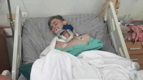 Kevin Lacey A photo of a man in a hospital bed. He is in a bed and he has medical machines attached to him. 