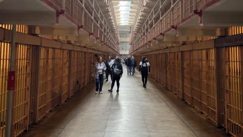 A group of tourists in sweats and sneakers strolls the long spare corridor in Alcatraz, with empty jail cells on either side. The only light shines in from skylights overhead