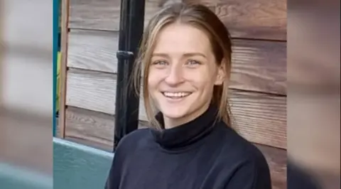 A young woman with light brown hair in a ponytail, wearing a black turtle neck and blue jeans sits on a bench. She is smiling at the camera.
