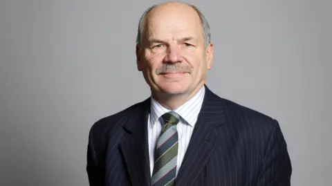 UK Parliament Earl Attlee wearing a suit and tie for his official House of Lords portrait in 2019.