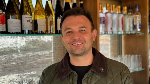 Jonathan Dryden is standing at the bar, bottles behind him, and smiling at the camera. He has short brown hair and stubble.