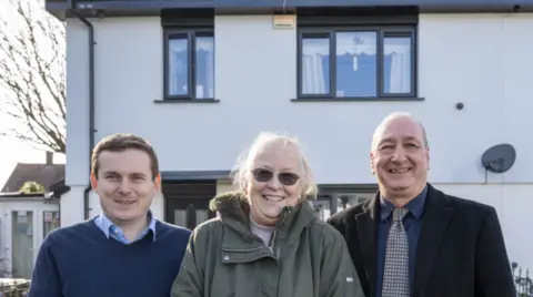 A man on the left is in a blue jumper, the woman in the middle is wearing a green coat and the man on the right is wearing a dark jacket, a tie and a shirt. The house behind them is white and they are looking at the camera.