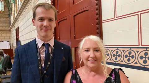 Man wearing a blue checked jacket and waistcoat with a white shirt and blue tie. He has a black lanyard round his neck. He has short blonde hair
A woman on his right is wearing a black and white dress. She has blonde hair that she is wearing up.