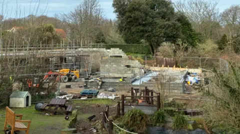 The construction site of the new Wildlife Hospital, with some brickwork done and construction equipment around, with a high fence around it and a small duck pond in front of it.