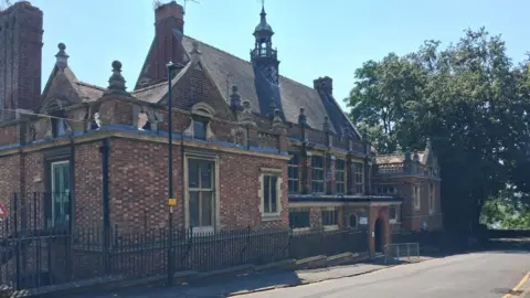 The ornate brick building has a turret with what looks like a bell tower. There are lots of stone pinnacles on the roof. The building is surrounded by railings and a tree grows next to it on the right.