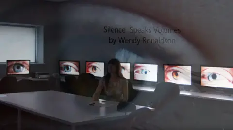 Wendy's Art A woman sits in a dark room surrounded by computers displaying close ups of eyes in varying colours. The lady has brown hair and a floral blouse and looks away from the camera, she sits at a white table with a blank piece of paper and a pen on. Over the top of the picture reads 'Silence Speaks Volumes by Wendy Ronaldson', and there is a blurry image of an eye barely visible.