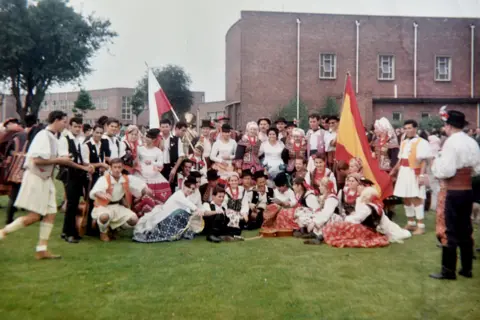 Walter Crinnion A big crowd of people sitting on grass. One person is holding a Spanish flag, the other is holding a Polish flag. Everyone is in traditional folk clothing.
