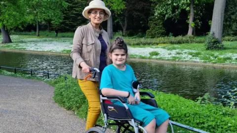Supplied Harbinder pushes Talvin in a wheelchair around a park, next to a river or pond. They both smile at the camera. She wears yellow jeggings and a beige jacket and sunhat. Talvin wears turquoise shorts and T-shirt. His hair is tied in a knot on top of his head.