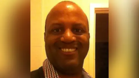 A head and shoulders selfie image of a man smiling at the camera, wearing a red white and blue checked shirt and dark jumper. He has brown eyes and has thinning dark hair