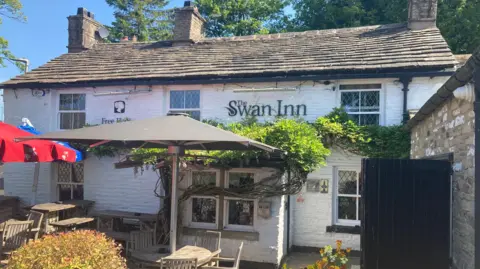 The outside of the traditional whitewashed Swan Inn pub with a small beer garden outside complete with parasols and tables and chairs.