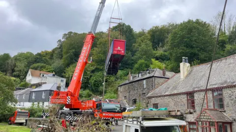 A red crane lifting a trailer that is wedged between a home and a wall. There are houses in the foreground and trees in the background.