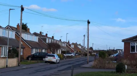 Grace Feeney A street with houses and cars parked on the road and on driveways, showing telegraph poles with wires coming off them.